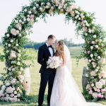 Newlyweds embracing under lush floral ceremony arch with countryside backdrop