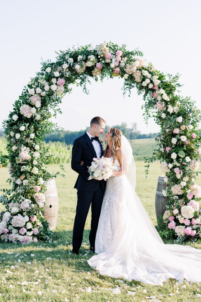 Newlyweds embracing under lush floral ceremony arch with countryside backdrop