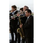 Horn section musicians in formal attire performing outdoors, featuring saxophone, trumpet, and trombone players