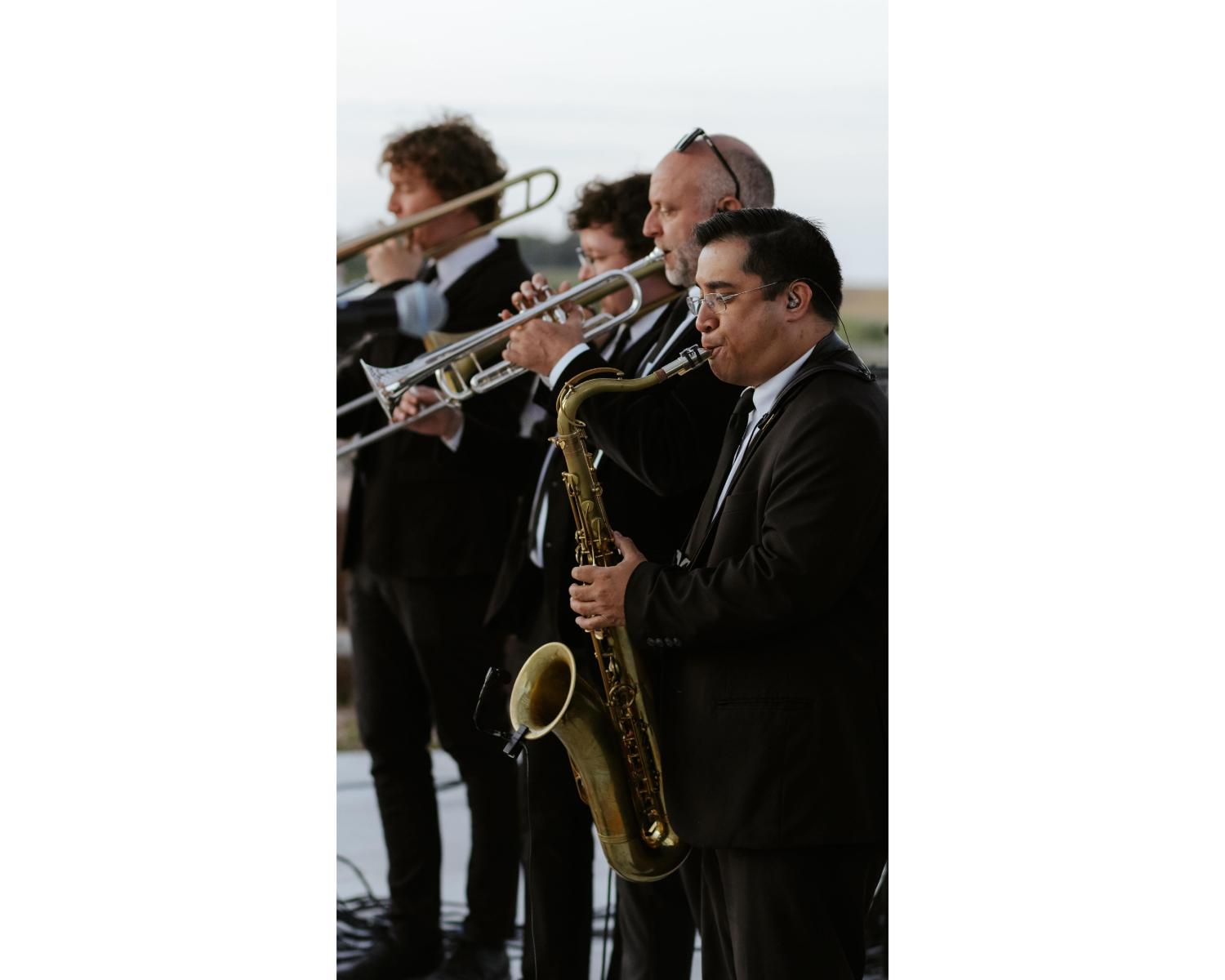 Horn section musicians in formal attire performing outdoors, featuring saxophone, trumpet, and trombone players