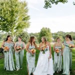Bride and bridesmaids in sage green dresses holding colorful bouquets on golf course lawn under trees