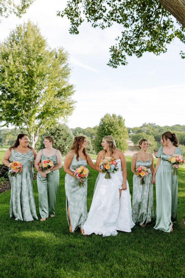 Bride and bridesmaids in sage green dresses holding colorful bouquets on golf course lawn under trees