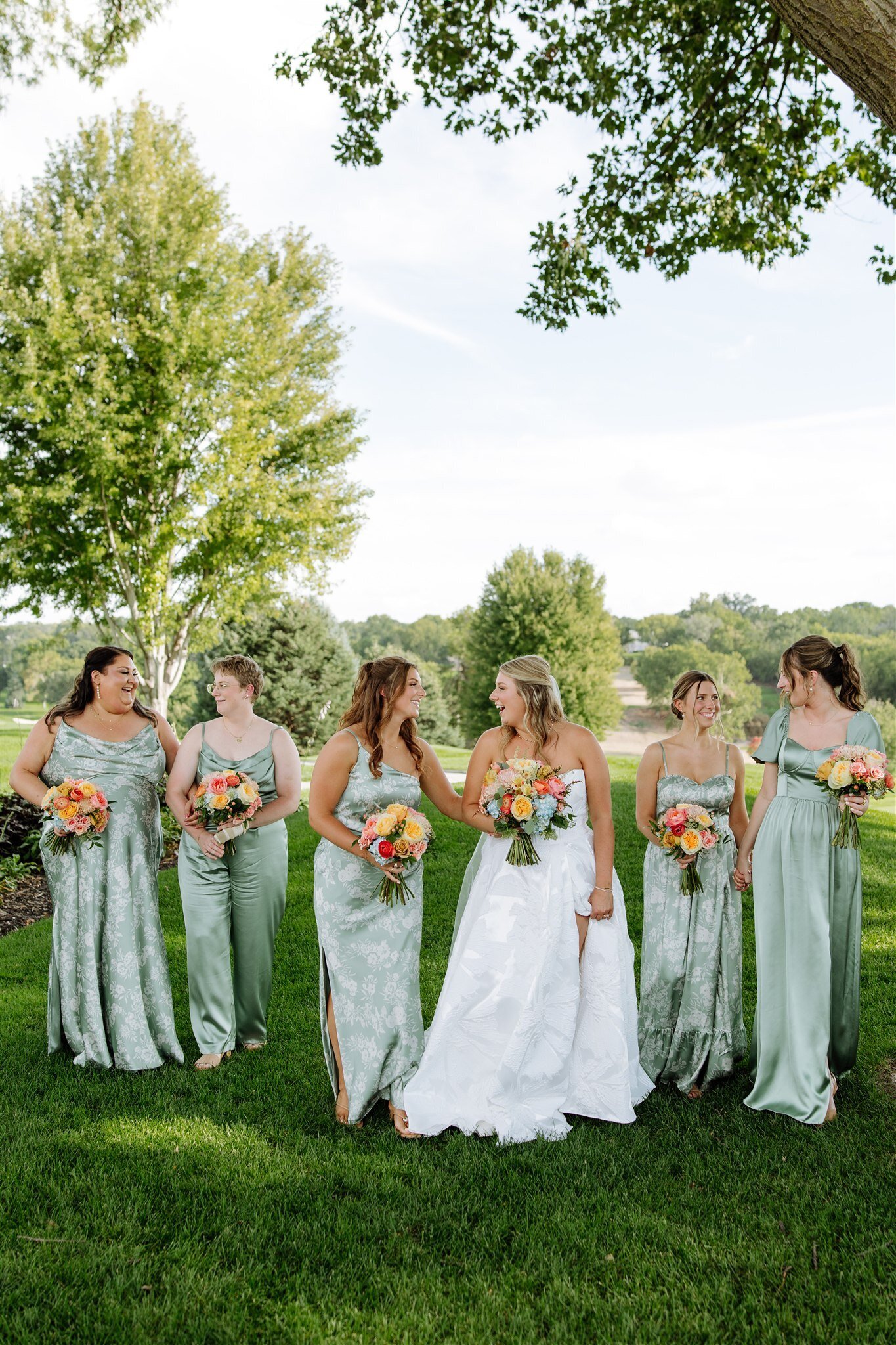 Bride and bridesmaids in sage green dresses holding colorful bouquets on golf course lawn under trees