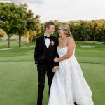 Newlyweds walk arm-in-arm across golf course green, groom in black tuxedo and bride in strapless white ball gown