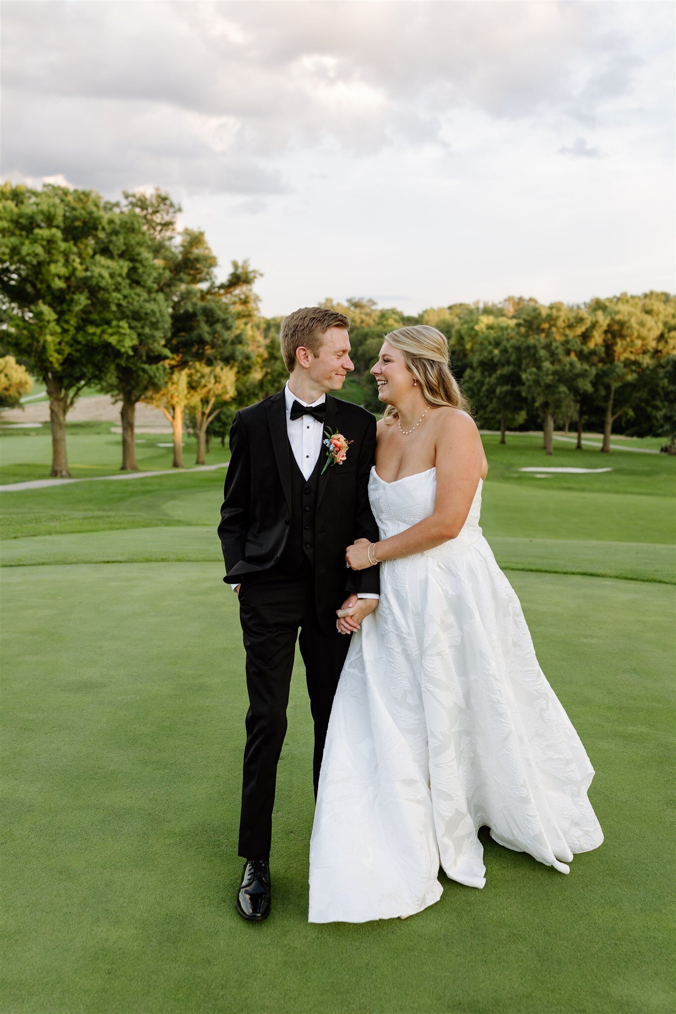 Newlyweds walk arm-in-arm across golf course green, groom in black tuxedo and bride in strapless white ball gown