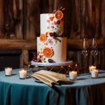 Two-tier white wedding cake decorated with dried orange slices, pressed flowers, and figs on a geometric cake stand surrounded by votive candles