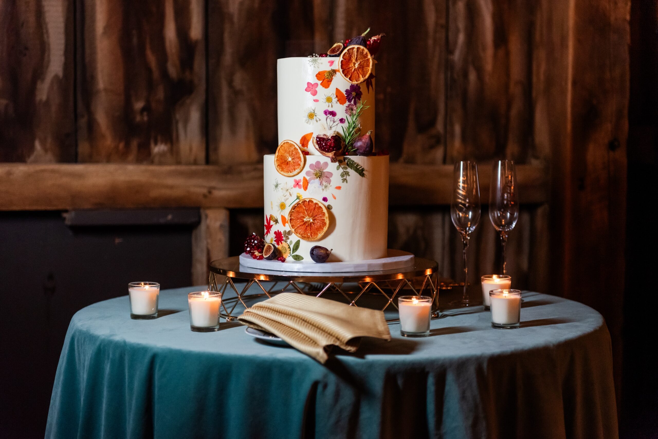 Two-tier white wedding cake decorated with dried orange slices, pressed flowers, and figs on a geometric cake stand surrounded by votive candles