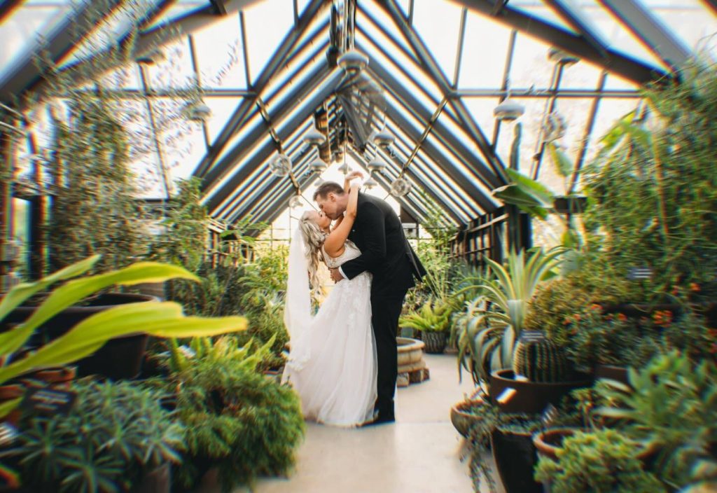 Bride and groom kissing in glass-enclosed greenhouse conservatory surrounded by tropical plants
