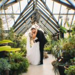 Bride and groom kissing in glass-enclosed greenhouse conservatory surrounded by tropical plants
