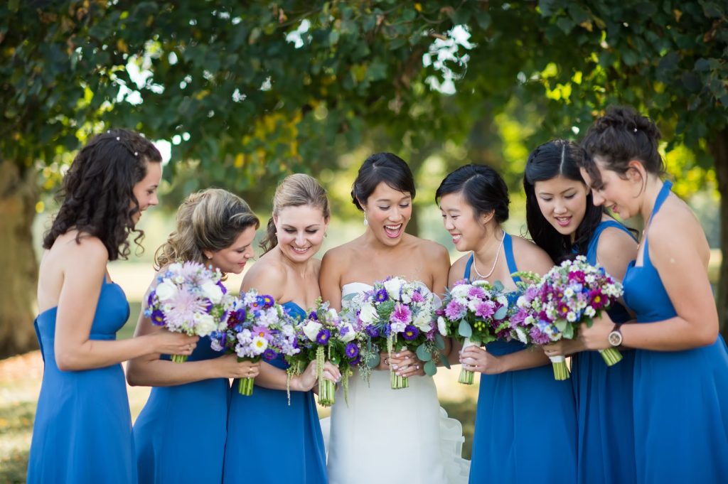 Bride and bridesmaids in blue dresses admiring purple and white bouquets in outdoor garden setting