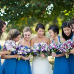 Bride and bridesmaids in blue dresses admiring purple and white bouquets in outdoor garden setting