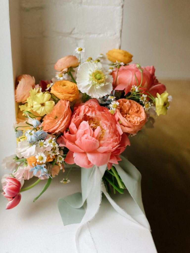 Colorful wedding bouquet with coral peonies, ranunculus, white poppies, daisies, and yellow blooms tied with trailing silk ribbons
