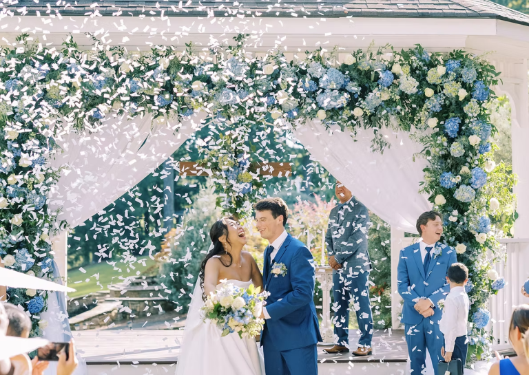 Newlyweds celebrating under blue hydrangea floral arch as confetti falls during outdoor ceremony