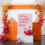 Rainbow gradient floral arch from yellow to pink roses framing welcome sign and orange bar display