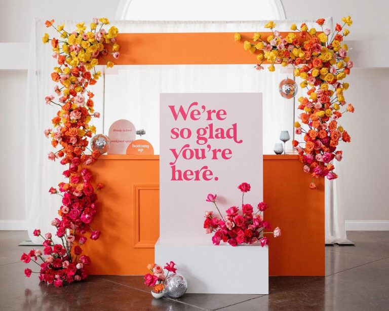 Rainbow gradient floral arch from yellow to pink roses framing welcome sign and orange bar display