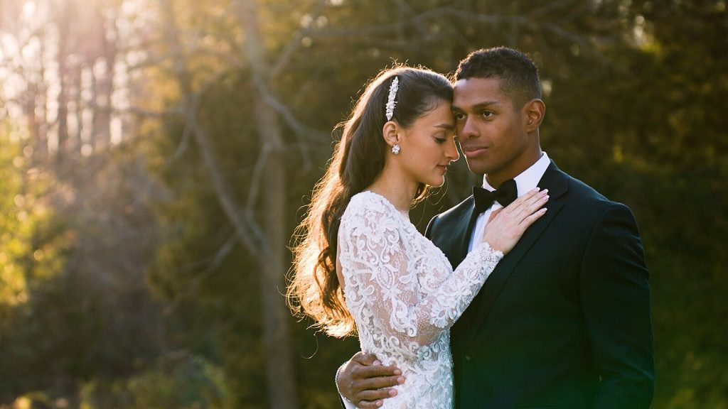 Bride in lace long-sleeve gown embracing groom in black tuxedo in golden hour sunlight with wooded background