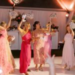 Wedding guests in pink formal dresses dancing with silver pom-poms under tent lighting