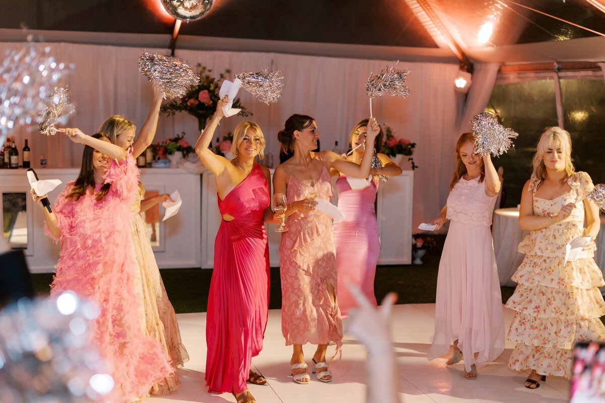 Wedding guests in pink formal dresses dancing with silver pom-poms under tent lighting