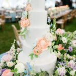 Five-tier white wedding cake adorned with cascading peach and pastel flowers in tented reception venue
