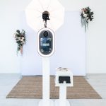 Modern white photo booth setup with ring light, umbrella lighting, and floral decorations