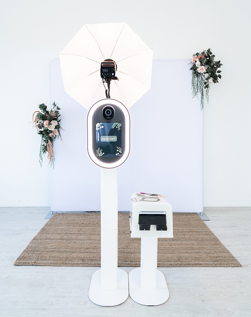 Modern white photo booth setup with ring light, umbrella lighting, and floral decorations