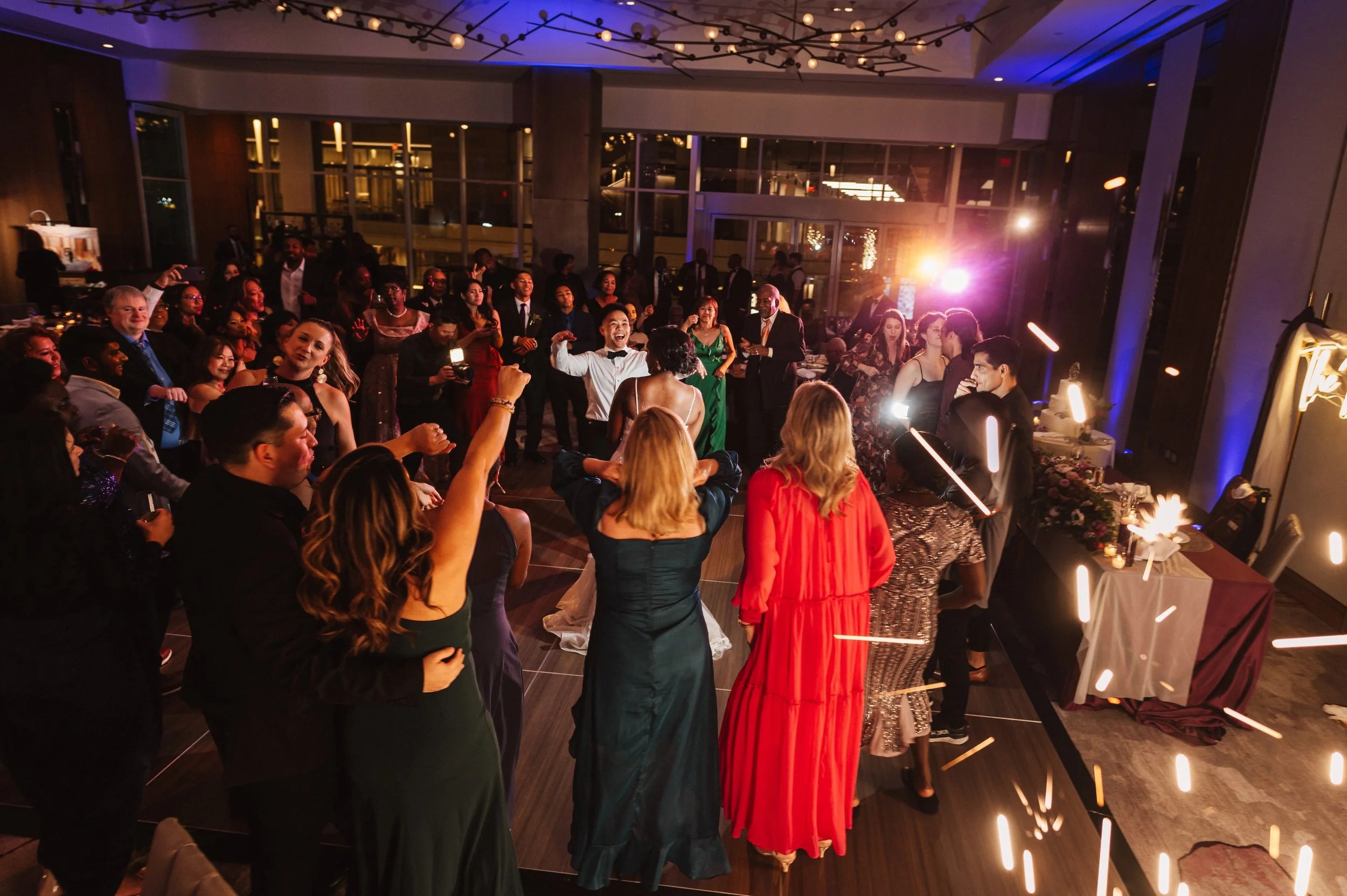 Wedding guests dancing at upscale reception venue with dramatic lighting and floor-to-ceiling windows