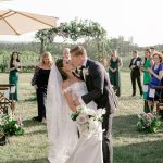 Newlyweds sharing first kiss as married couple during outdoor vineyard ceremony with floral arch