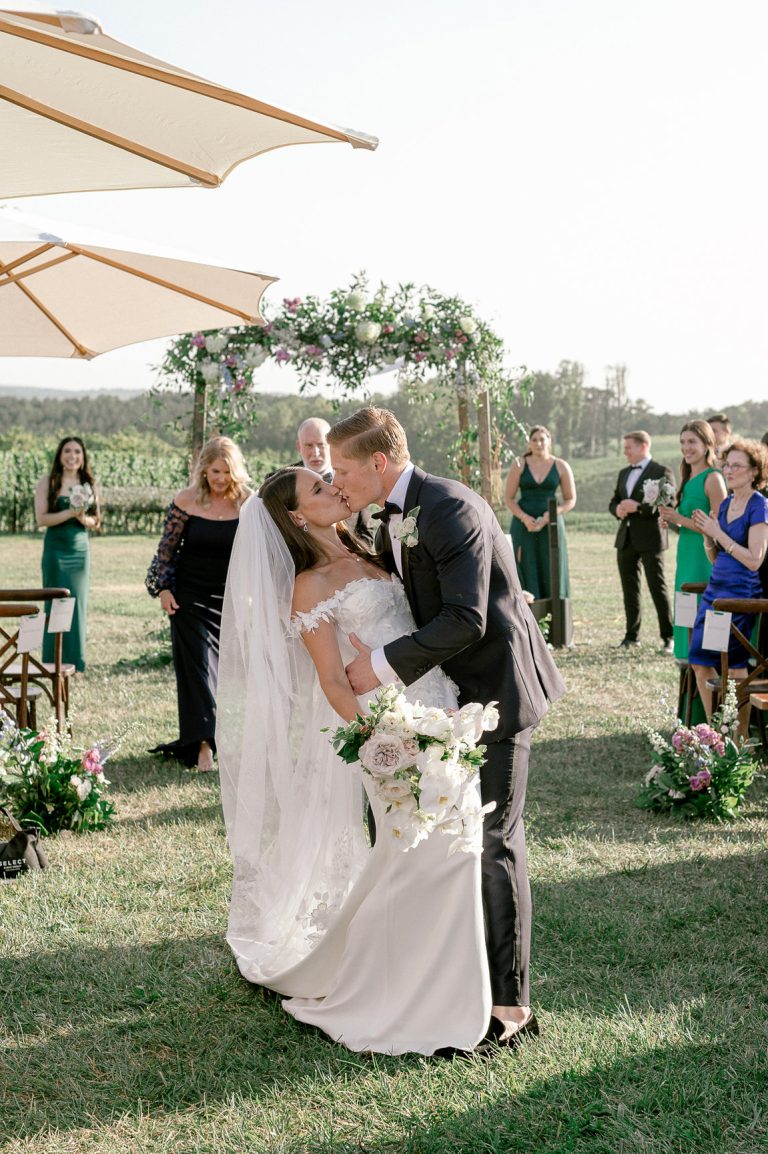 Newlyweds sharing first kiss as married couple during outdoor vineyard ceremony with floral arch