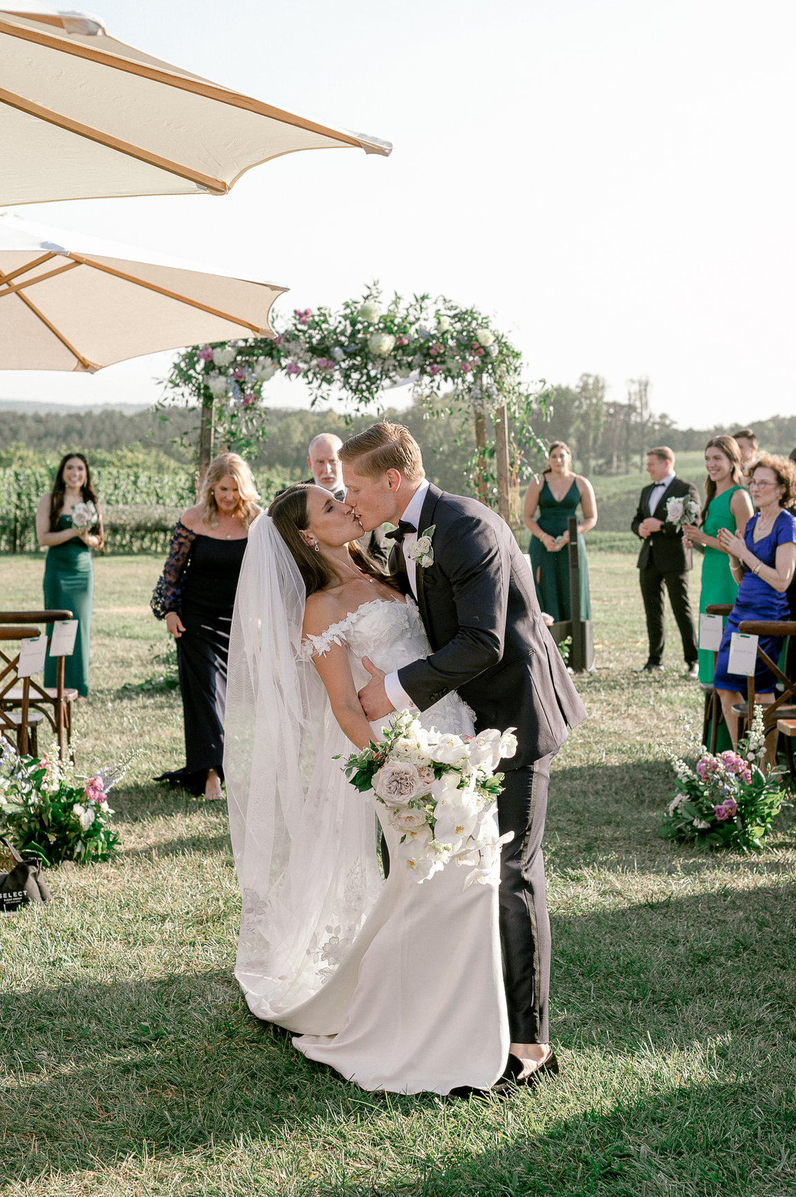 Newlyweds sharing first kiss as married couple during outdoor vineyard ceremony with floral arch