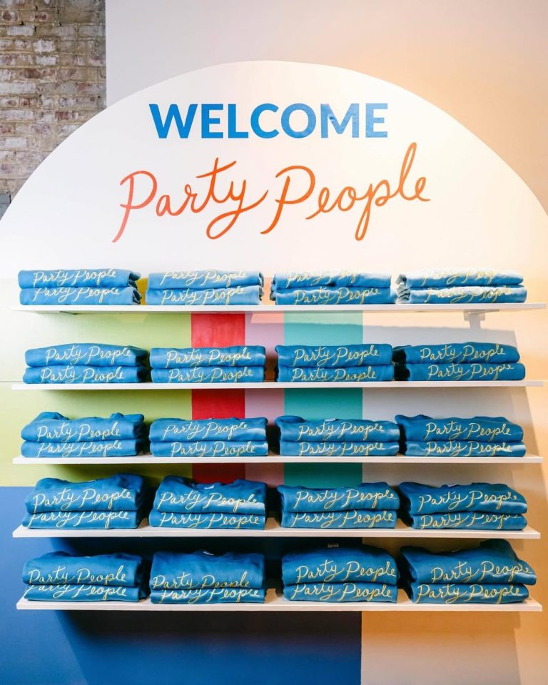White shelving display of blue "Party People" branded merchandise beneath circular welcome sign