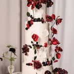 Four-tier white textured wedding cake decorated with red poppies, blackberries, grapes, and dark florals