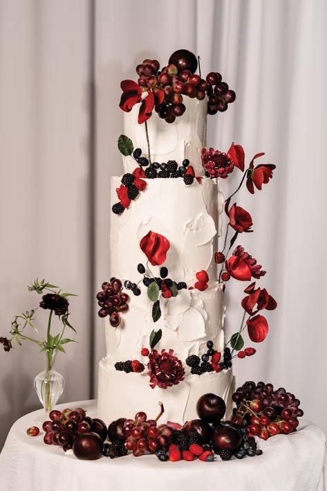 Four-tier white textured wedding cake decorated with red poppies, blackberries, grapes, and dark florals