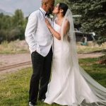 Bride in elegant white wedding dress with long cathedral veil kissing groom in white jacket outdoors with mountain backdrop