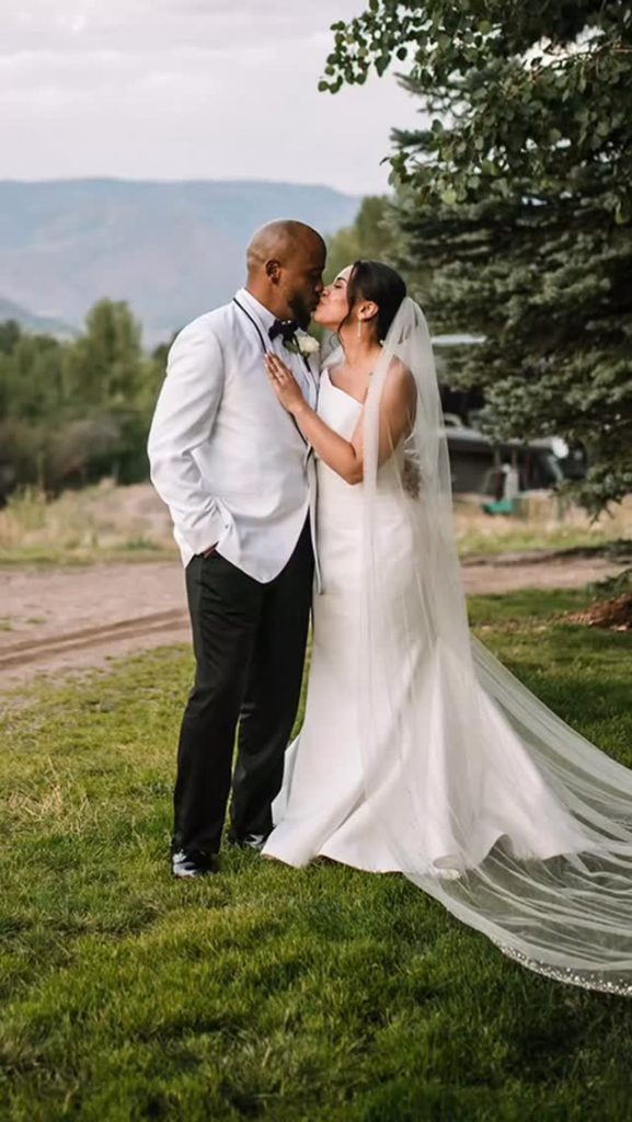 Bride in elegant white wedding dress with long cathedral veil kissing groom in white jacket outdoors with mountain backdrop