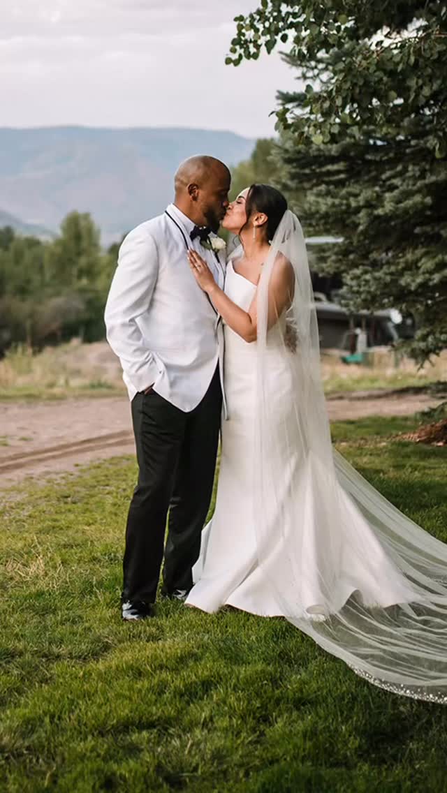 Bride in elegant white wedding dress with long cathedral veil kissing groom in white jacket outdoors with mountain backdrop