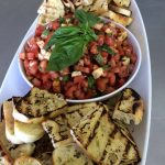 Fresh bruschetta with diced tomatoes, mozzarella, and basil surrounded by grilled bread slices on white platter