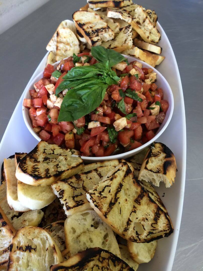 Fresh bruschetta with diced tomatoes, mozzarella, and basil surrounded by grilled bread slices on white platter
