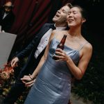 Joyful couple embracing under red awning at outdoor celebration with wedding guest in background