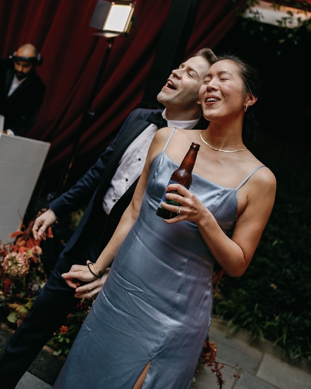 Joyful couple embracing under red awning at outdoor celebration with wedding guest in background