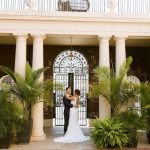 Bride and groom embracing under white columned portico with tropical plants and ornate iron gate