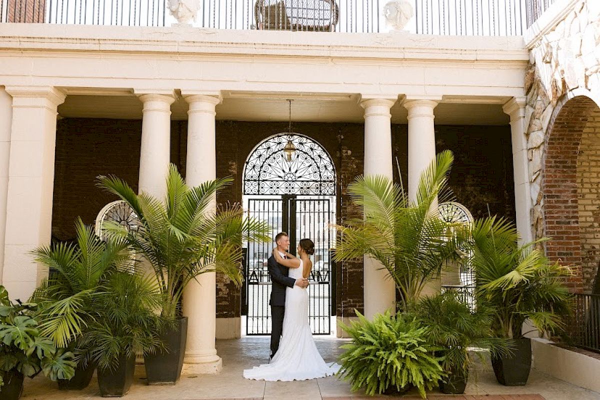 Bride and groom embracing under white columned portico with tropical plants and ornate iron gate