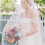 Bride in off-shoulder gown and veil holding colorful bouquet outdoors