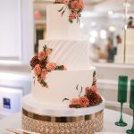 Three-tier white wedding cake decorated with coral and burgundy sugar roses and dog topper on crystal cake stand