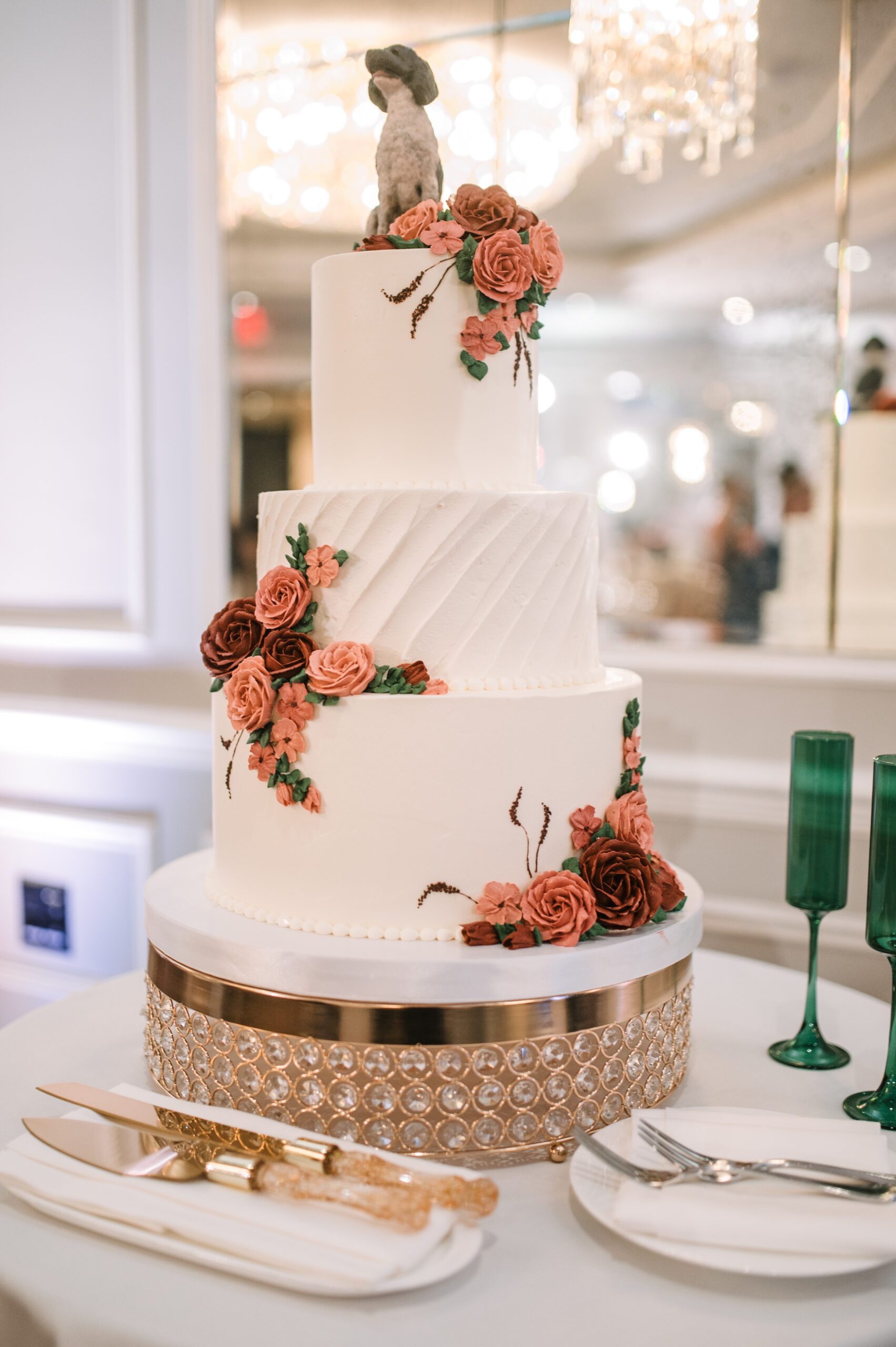 Three-tier white wedding cake decorated with coral and burgundy sugar roses and dog topper on crystal cake stand