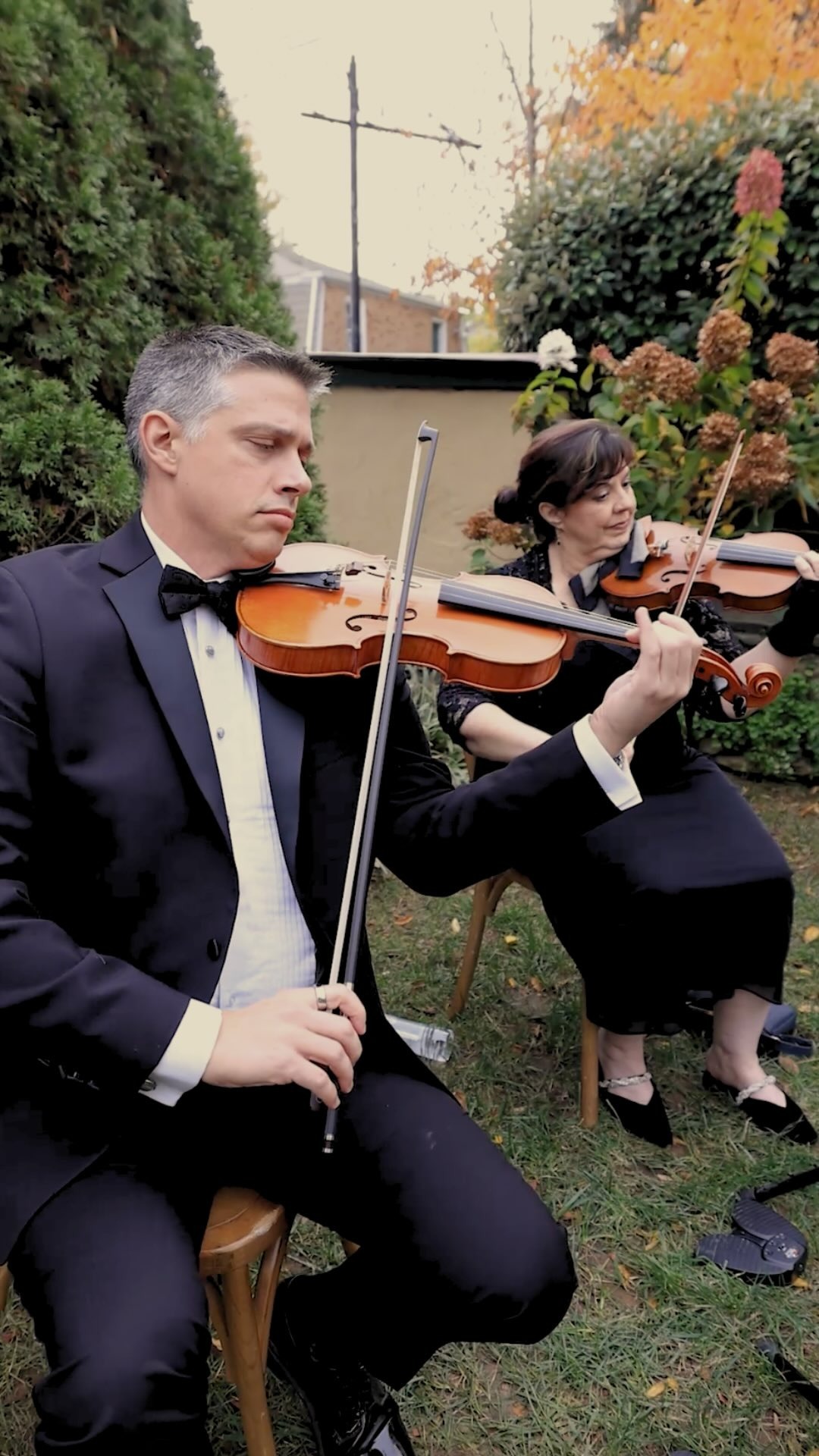 Two formally dressed violinists performing outdoors in an autumn garden setting