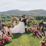 Bride and groom sharing first kiss during outdoor ceremony with pink floral arch and mountain views