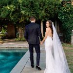 Bride in lace gown with flowing veil looks back at camera while holding groom's hand by poolside
