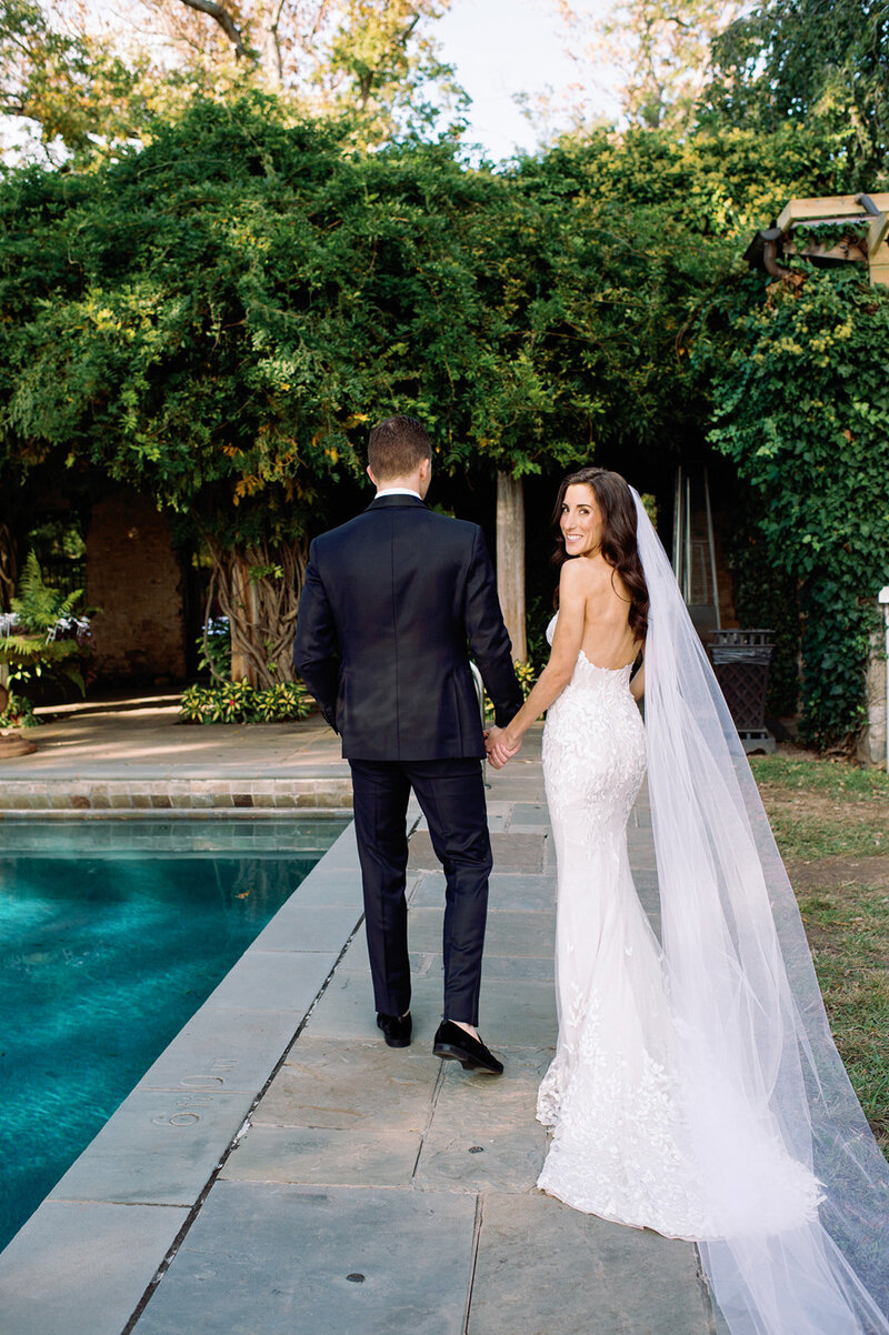 Bride in lace gown with flowing veil looks back at camera while holding groom's hand by poolside