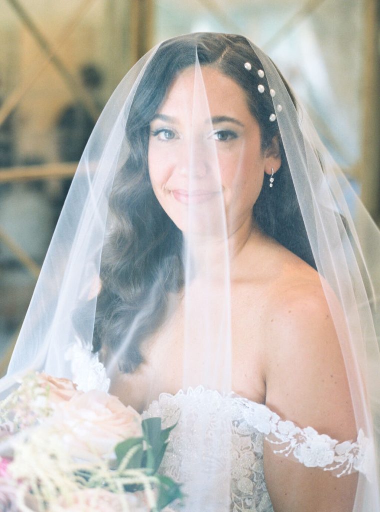 Bride wearing pearl-adorned veil with natural makeup and loose waves holding bouquet