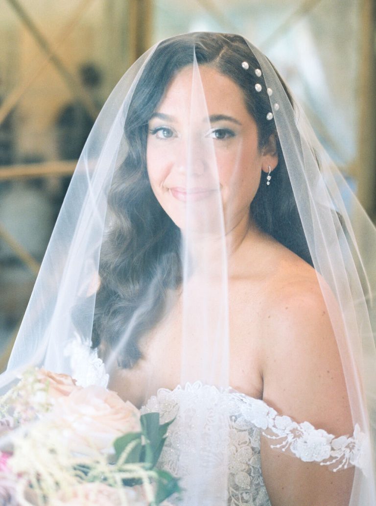 Bride wearing pearl-adorned veil with natural makeup and loose waves holding bouquet